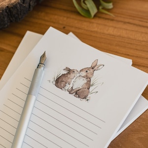 May include: Lined stationery featuring a watercolour illustration of two brown rabbits. A silver fountain pen rests on the top sheet. The stationery is on a wooden surface.