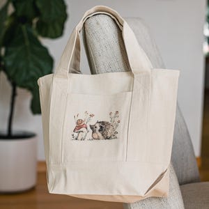 May include: A cream-coloured tote bag featuring a charming illustration of a snail and a hedgehog amongst flowers. The bag has a front pocket and strong handles. Made from durable canvas, it's ideal for carrying shopping or everyday items.