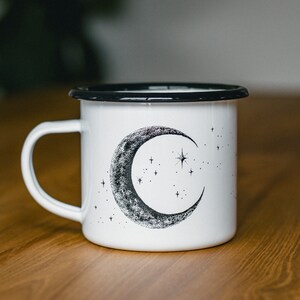 May include: White enamel mug with a black rim and a black and white illustration of a crescent moon and stars.