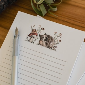 Puede incluir: Un bloc de papel de carta rayado con una ilustración caprichosa de un erizo y un caracol. Una pluma estilográfica plateada descansa sobre la hoja superior. La ilustración presenta una seta y flores silvestres. El papel es blanco con líneas grises.