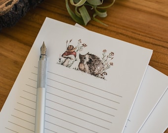 May include: A stack of lined stationery with a whimsical illustration of a hedgehog and snail. A silver fountain pen rests on the top sheet. The illustration features a mushroom and wildflowers. The paper is white with gray lines.