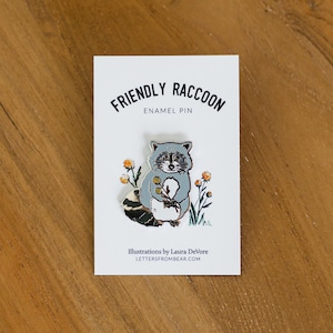 Puede incluir: Un pin de esmalte "Friendly Raccoon" en una tarjeta blanca. El pin presenta un mapache gris y blanco sosteniendo flores, con el texto "Friendly Raccoon" arriba y "Enamel Pin" abajo. La tarjeta está sobre una superficie de madera.