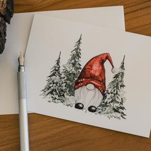 May include: A watercolor Christmas card featuring a gnome wearing a red hat and surrounded by evergreen trees. The card is on a white envelope, with a silver fountain pen in the foreground. The overall theme is holiday greetings.