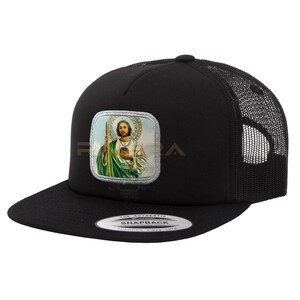 May include: Black trucker hat with a mesh back and a flat brim. The front features a square patch with an image of a religious figure. The hat has a snapback closure and a white tag with the text "SNAPBACK".
