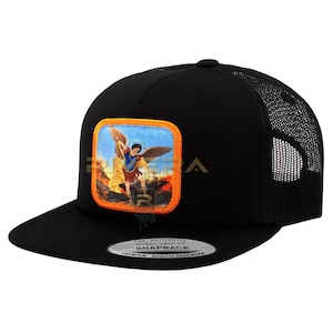May include: Black trucker hat with a mesh back and a flat brim. The front features a square patch with a colorful illustration of an angel. The hat has a snapback closure and a curved brim. The text on the brim reads "SNAPBACK".