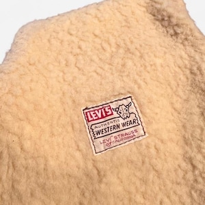 May include: A cream-colored sherpa fleece fabric featuring a rectangular white fabric label stitched onto the center. The label displays the red text LEVI'S, a graphic of a bull skull, and the black text AUTHENTIC WESTERN WEAR LEVI STRAUSS OF CALIFORNIA.