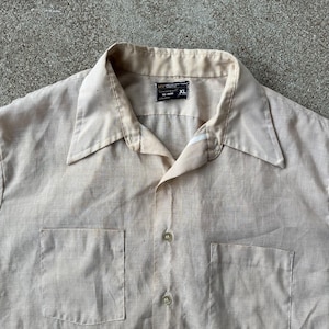May include: A light tan button-down shirt with two front pockets. The shirt has a pointed collar and a label that reads "MW" and "XL". The shirt is made of a textured fabric and has a vintage look.