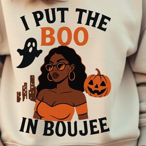 May include: Beige sweatshirt with the text "I PUT THE BOO IN BOUJEE". The graphic includes a black ghost, a woman wearing sunglasses, and an orange jack-o'-lantern. The woman is wearing an orange top. The sweatshirt is a Halloween-themed design.