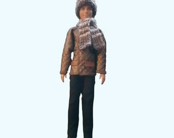 May include: A doll wearing a gray and white faux fur hat, a matching gray and white knit scarf, and a brown quilted jacket with a textured pattern. The doll is also dressed in solid black trousers and brown shoes, standing in an upright position.