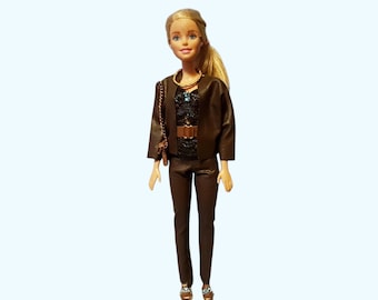 May include: A blonde doll wearing a dark brown jacket, matching trousers, and a black textured top with a gold-colored belt. The doll features a gold-colored necklace, a small brown shoulder bag with a gold chain, and metallic gold-colored shoes.