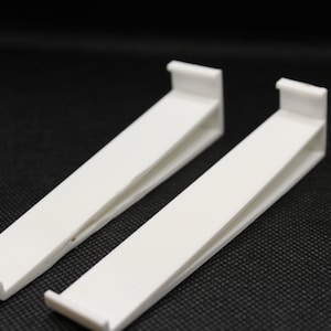 Puede incluir: Dos soportes de estante de plástico blanco. Los soportes están inclinados y tienen un diseño simple.