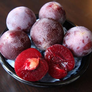 May include: A close-up of a black bowl filled with plums. Some plums are whole, with a deep purple skin and a white, speckled coating. Two plums are cut in half, revealing a vibrant red flesh and a single pit.