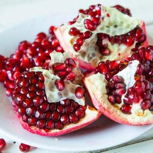 May include: A white plate holds three sections of a pomegranate, showcasing vibrant red seeds. The fruit's rind is a mix of red and cream, with the seeds clustered inside. Scattered seeds add to the fresh, natural presentation.