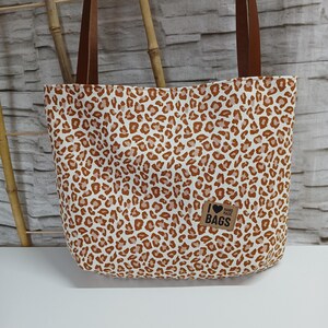Könnte beinhalten: Eine braune und weiße Leopardenmuster-Tragetasche mit braunen Lederriemen. Die Tasche hat einen kleinen Aufnäher auf der Vorderseite, auf dem "Ich liebe handgemachte Taschen" steht.