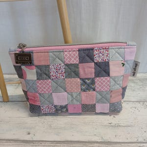 Könnte beinhalten: Eine gesteppte Kosmetiktasche aus Patchwork in Rosa- und Grautönen mit Reißverschluss und kleinem Metallanhänger. Die Tasche besteht aus verschiedenen gemusterten Stoffen, darunter Blumen- und geometrische Designs. Das Etikett lautet "made with magic".