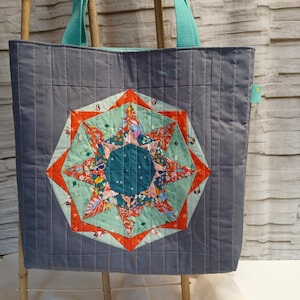 Könnte beinhalten: Graue, gesteppte Tragetasche mit einem zentralen Sternmuster. Der Stern hat eine türkisfarbene Mitte, umgeben von orangefarbenen und floral gemusterten Segmenten. Türkisfarbene Griffe vervollständigen das Design der Tasche.