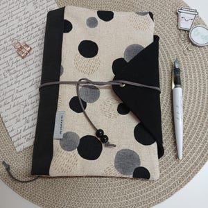 Könnte beinhalten: Ein Stoff-Journal-Cover mit beigem Hintergrund und großen schwarzen und grauen Polka-Dots. Die Hülle hat einen schwarzen Klappenverschluss und wird mit einer grauen Kordel gebunden. Ein weißer Stift und dekorative Büroklammern sind ebenfalls zu sehen.