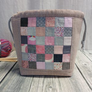 Könnte beinhalten: Ein beigefarbener Beutel mit Kordelzug und einer Patchwork-Vorderseite. Der Patchwork besteht aus kleinen Quadraten in verschiedenen Rosa-, Grau- und gemusterten Stoffen. Der Beutel hat einen grauen Kordelzugverschluss.