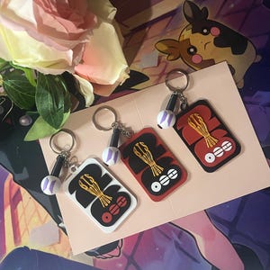 May include: Three rectangular keychains featuring a black and red design with a gold-colored graphic and three white circles. Each keychain includes a small, round, white charm with a purple logo attached to a silver metal ring and chain.