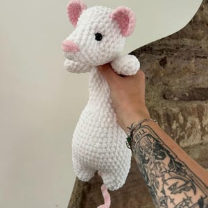 Könnte beinhalten: Eine handgefertigte weiße Häkelmaus mit rosa Ohren, Nase und Schwanz. Das Stofftier hat schwarze Knopfaugen und wird von einer Hand gehalten. Das weiche, strukturierte Garn verleiht der Maus ein kuscheliges Aussehen. Geeignet für Kinder oder als Dekoration.