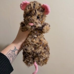 May include: A handmade plush toy mouse with a brown and tan spotted body, pink ears, and a pink tail. The mouse has black eyes and a pink tongue sticking out. The toy is made of soft, fuzzy material.