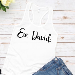May include: White tank top with the text "Ew, David." printed in black cursive font. The tank top is laying on a light wood surface with a pair of denim shorts.