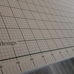May include: An off-white cutting mat featuring a grid pattern and measurements in centimetres. The mat is marked with the text "aqua by design". Black grid lines and measurement markings are visible along the bottom edge.