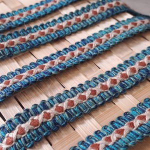 May include: Close-up of decorative trim with a woven design. The trim features a repeating pattern of blue, teal, and brown colours. The trim is placed on a woven bamboo surface.