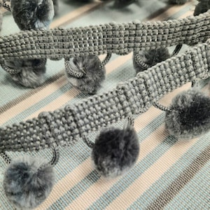 May include: Grey trim with fuzzy pom poms. The trim is woven and has a textured look. The pom poms are soft and fluffy.