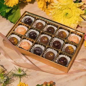 K&ouml;nnte beinhalten: Eine goldene Schachtel gef&uuml;llt mit verschiedenen Pralinen, jede in einer Papierform. Die Pralinen haben verschiedene Farben und Designs, umgeben von gelben und orangen Blumen, mit einem rosa Band. Ein Geschenk f&uuml;r jeden Anlass.