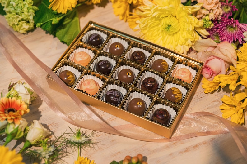K&ouml;nnte beinhalten: Eine goldene Schachtel gef&uuml;llt mit verschiedenen Pralinen, jede in einer Papierform. Die Pralinen haben verschiedene Farben und Designs, umgeben von gelben und orangen Blumen, mit einem rosa Band. Ein Geschenk f&uuml;r jeden Anlass.