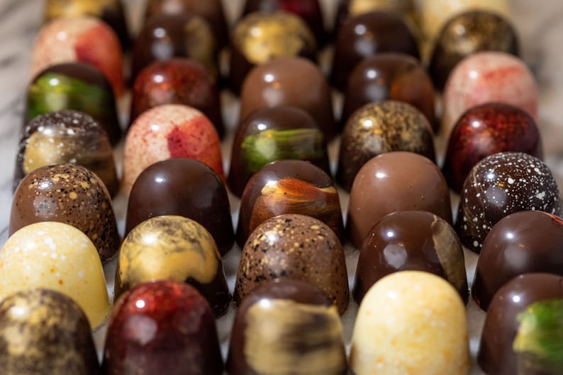 K&ouml;nnte beinhalten: Eine Auswahl handgefertigter Pralinen mit verschiedenen Farben und Texturen. Die Pralinen sind in ordentlichen Reihen angeordnet und zeigen eine Vielzahl von Geschmacksrichtungen und Designs, darunter gesprenkelte, gewirbelte und einfarbige Farben. Das Bild hebt die Details jedes St&uuml;cks hervor.