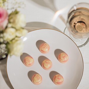 K&ouml;nnte beinhalten: Ein wei&szlig;er Teller mit Goldrand enth&auml;lt sechs rosenf&ouml;rmige Pralinen. Ein Glas rosa Fl&uuml;ssigkeit steht daneben, und ein Blumenstrau&szlig; ist im Hintergrund. Die Pralinen haben ein rosa-cremefarbenes Wirbeldesign.