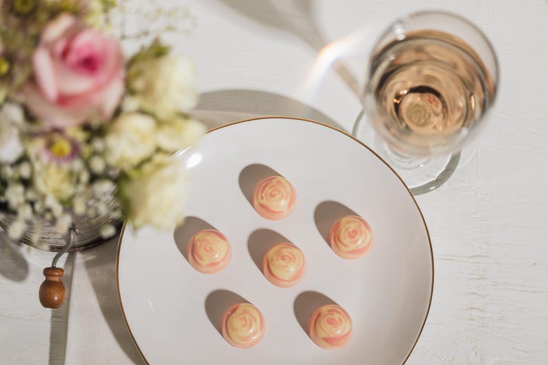 K&ouml;nnte beinhalten: Ein wei&szlig;er Teller mit Goldrand enth&auml;lt sechs rosenf&ouml;rmige Pralinen. Ein Glas rosa Fl&uuml;ssigkeit steht daneben, und ein Blumenstrau&szlig; ist im Hintergrund. Die Pralinen haben ein rosa-cremefarbenes Wirbeldesign.