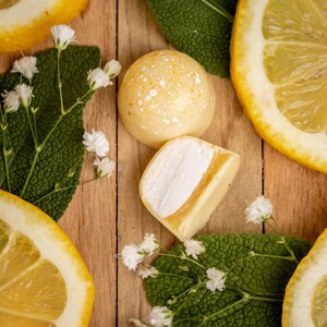 K&ouml;nnte beinhalten: Draufsicht auf eine Holzoberfl&auml;che mit Zitronenscheiben, gr&uuml;nen Bl&auml;ttern und kleinen wei&szlig;en Bl&uuml;ten. Zwei gelbe S&uuml;&szlig;igkeiten, eine ganz und eine geschnittene, sind ebenfalls vorhanden. Die S&uuml;&szlig;igkeiten haben eine wei&szlig;e F&uuml;llung und ein gesprenkeltes Muster.