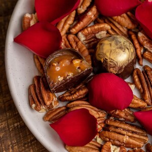 K&ouml;nnte beinhalten: Ein wei&szlig;er Teller mit Pekann&uuml;ssen, roten Rosenbl&auml;ttern und zwei Pralinen. Eine Praline ist halbiert und zeigt eine Karamellf&uuml;llung. Die andere hat eine goldfarbene Oberseite.