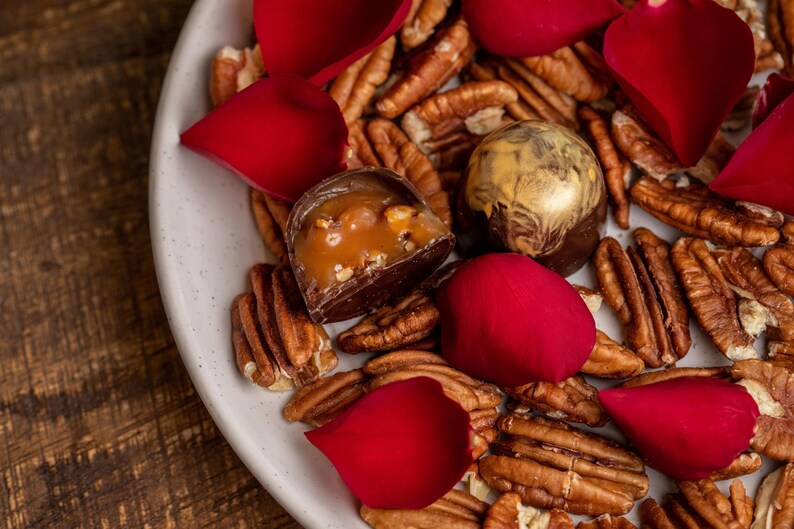 K&ouml;nnte beinhalten: Ein wei&szlig;er Teller mit Pekann&uuml;ssen, roten Rosenbl&auml;ttern und zwei Pralinen. Eine Praline ist halbiert und zeigt eine Karamellf&uuml;llung. Die andere hat eine goldfarbene Oberseite.