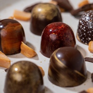 K&ouml;nnte beinhalten: Eine Auswahl an Pralinen mit verschiedenen Designs und Farben, darunter Dunkelbraun, Gold und Rot. Einige Pralinen haben wei&szlig;e Sprenkel. Karamellst&uuml;cke und Schokoladenst&uuml;cke sind auf einem wei&szlig;en Teller verstreut.