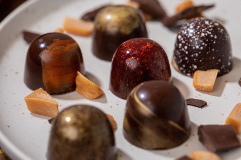 K&ouml;nnte beinhalten: Eine Auswahl an Pralinen mit verschiedenen Designs und Farben, darunter Dunkelbraun, Gold und Rot. Einige Pralinen haben wei&szlig;e Sprenkel. Karamellst&uuml;cke und Schokoladenst&uuml;cke sind auf einem wei&szlig;en Teller verstreut.