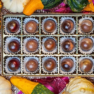May include: A rectangular gold box containing fifteen assorted round chocolates, each nestled in a white paper liner. The chocolates feature various brown, bronze, and dark red glossy finishes. The box is surrounded by several decorative gourds.
