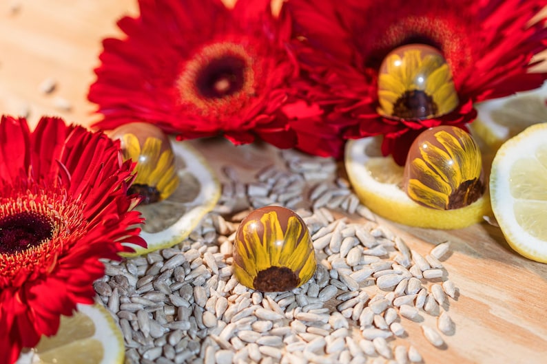 K&ouml;nnte beinhalten: Nahaufnahme von sonnenblumenf&ouml;rmigen Pralinen mit gelben Bl&uuml;tenbl&auml;ttern, die auf Sonnenblumenkernen liegen. Rote Gerbera und Zitronenscheiben erg&auml;nzen die Szene und erzeugen ein farbenfrohes Arrangement.