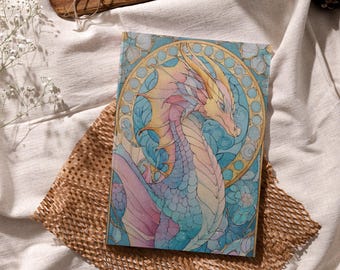 May include: A hardcover journal featuring a colorful illustration of a dragon with a stained-glass style design. The dragon has a pink, yellow, and blue color palette, with a gold border. The journal is resting on a brown paper.