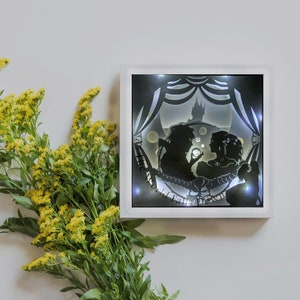 May include: A square, white-framed light box featuring a silhouette scene of two figures in a romantic setting. The black and white image includes a castle and decorative elements. Yellow flowers are in the foreground.