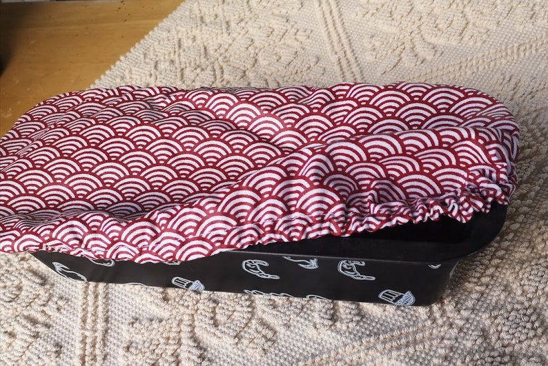 May include: A red and white patterned fabric loaf pan cover. The cover is made of a soft, stretchy fabric and has a scalloped edge. The cover is sitting on top of a black loaf pan.
