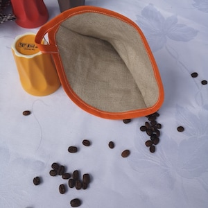 以下が含まれることがあります： オレンジ色の縁取りが付いた薄い茶色のリネン製のコーヒーウォーマーのクローズアップ。ウォーマーは白いテーブルクロスの上に置かれ、周りにコーヒー豆が散らばっています。背景にはオレンジ色と黄色のセラミック製のマグカップが2つあります。