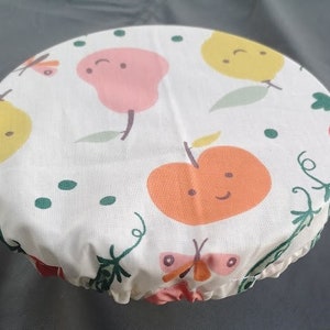 K&ouml;nnte beinhalten: Wei&szlig;er Stoff-Sch&uuml;sseldeckel mit einem bunten Obst- und Schmetterlingsdesign. Der Deckel zeigt eine Birne, einen Apfel und einen Schmetterling mit einem gr&uuml;nen Stiel und Bl&auml;ttern.