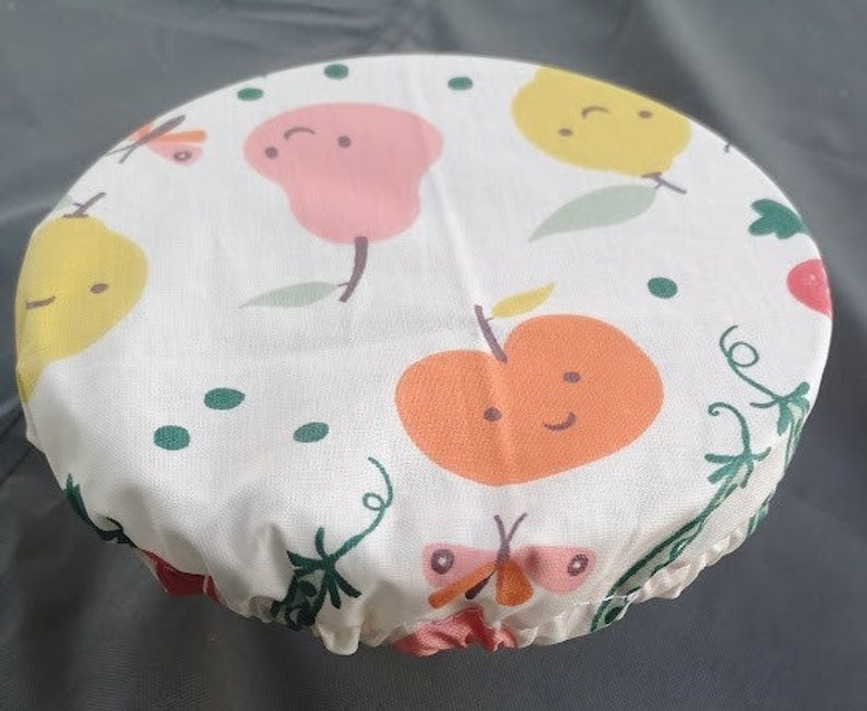 K&ouml;nnte beinhalten: Wei&szlig;er Stoff-Sch&uuml;sseldeckel mit einem bunten Obst- und Schmetterlingsdesign. Der Deckel zeigt eine Birne, einen Apfel und einen Schmetterling mit einem gr&uuml;nen Stiel und Bl&auml;ttern.