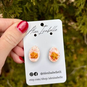 May include: A pair of novelty earrings shaped like deviled eggs, with a white and pink speckled egg white and a yellow yolk. The earrings are displayed on a white card with the text "Alex Isabelle" and social media handles.