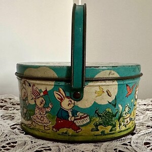 Puede incluir: Una fiambrera vintage de forma ovalada con un asa turquesa. La fiambrera presenta una ilustraci&oacute;n caprichosa de conejos, ranas y p&aacute;jaros sobre un fondo azul y blanco. El texto "Happy Bunny" es visible en la parte inferior de la fiambrera.