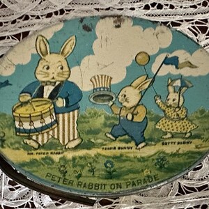 Puede incluir: Una fiambrera de metal vintage con una ilustraci&oacute;n colorida de Peter Rabbit y otros conejos en un desfile. Los conejos est&aacute;n vestidos con atuendos patri&oacute;ticos, uno tocando un tambor y otro sosteniendo un globo. El texto "Peter Rabbit on Parade" est&aacute; impreso en la parte inferior de la fiambrera.